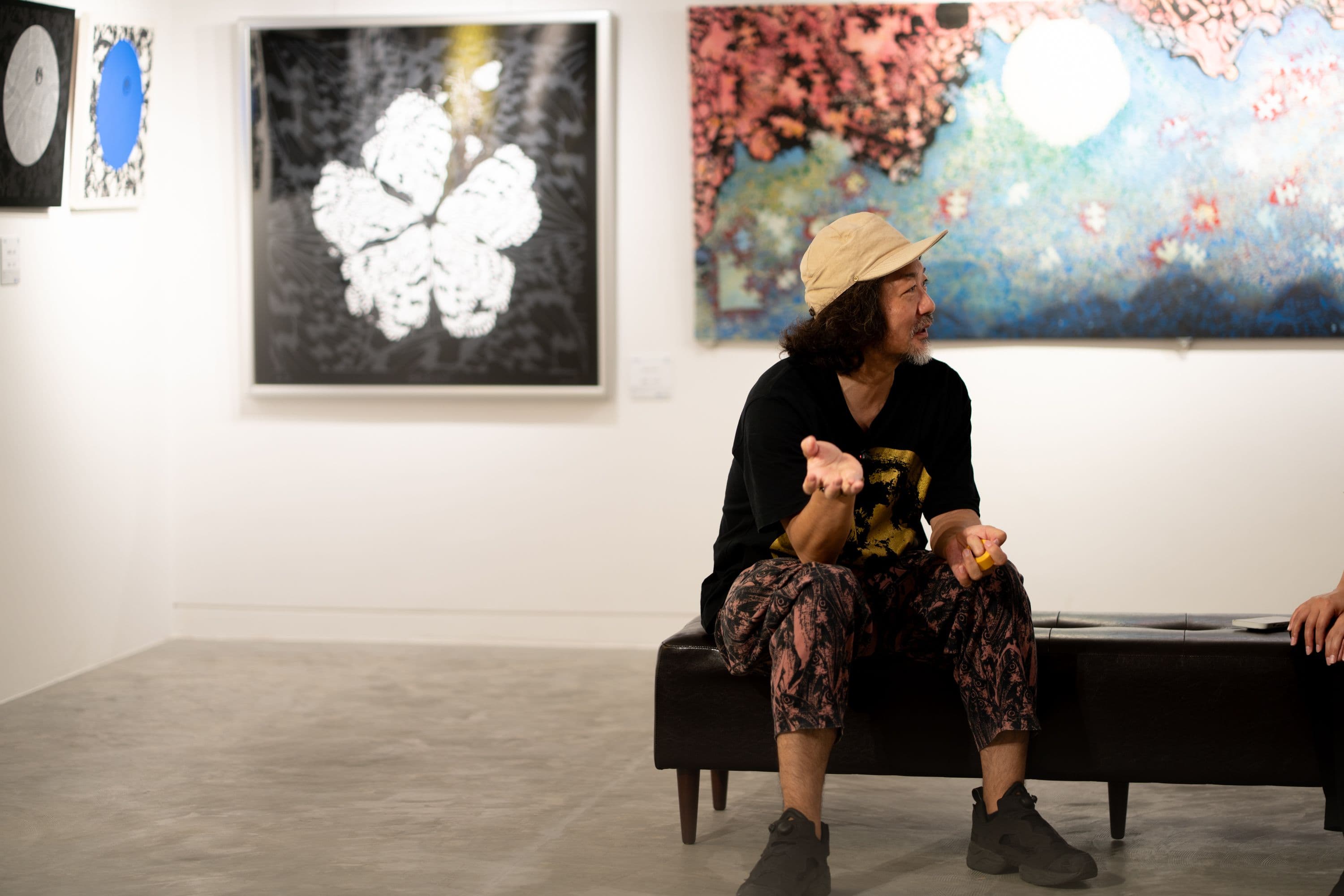 花の作品と大きな絵画の前で、ベンチに座って話す池城安武さん