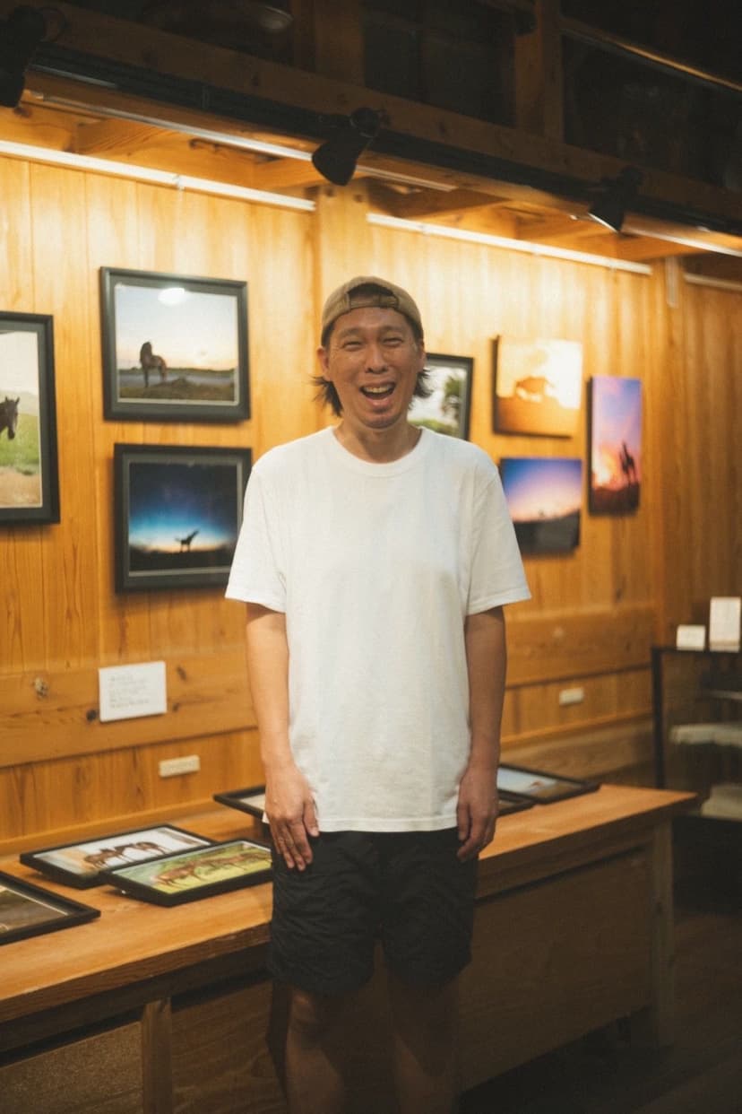木の壁に写真作品が並ぶギャラリーで、笑顔で立つ能津敦司さん